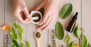 Pessoa mexendo substâncias medicamentos para formação de medicamentos homeopáticos em uma mesa de madeira ao lado de folha de planta, frasco de vidro e colheres.